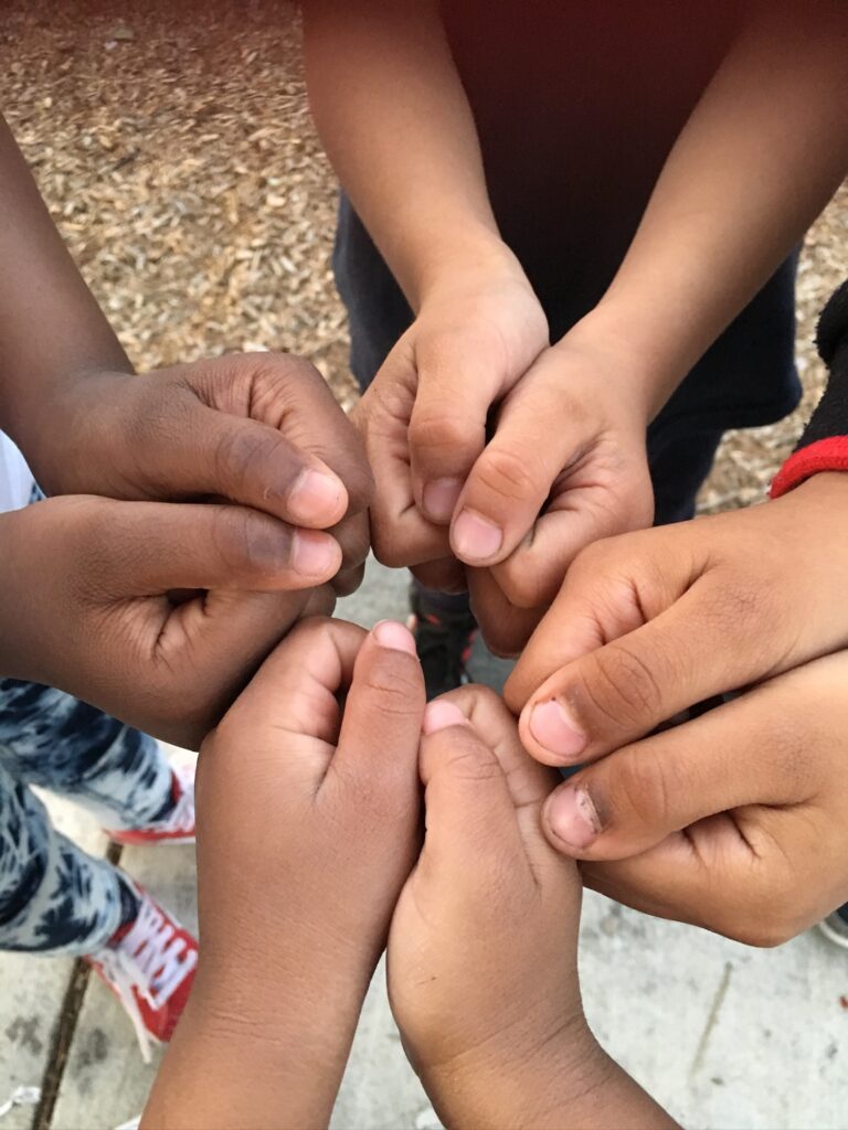 Children putting their hands together, showing tops of thumbs , like a fist pound in a circle together.