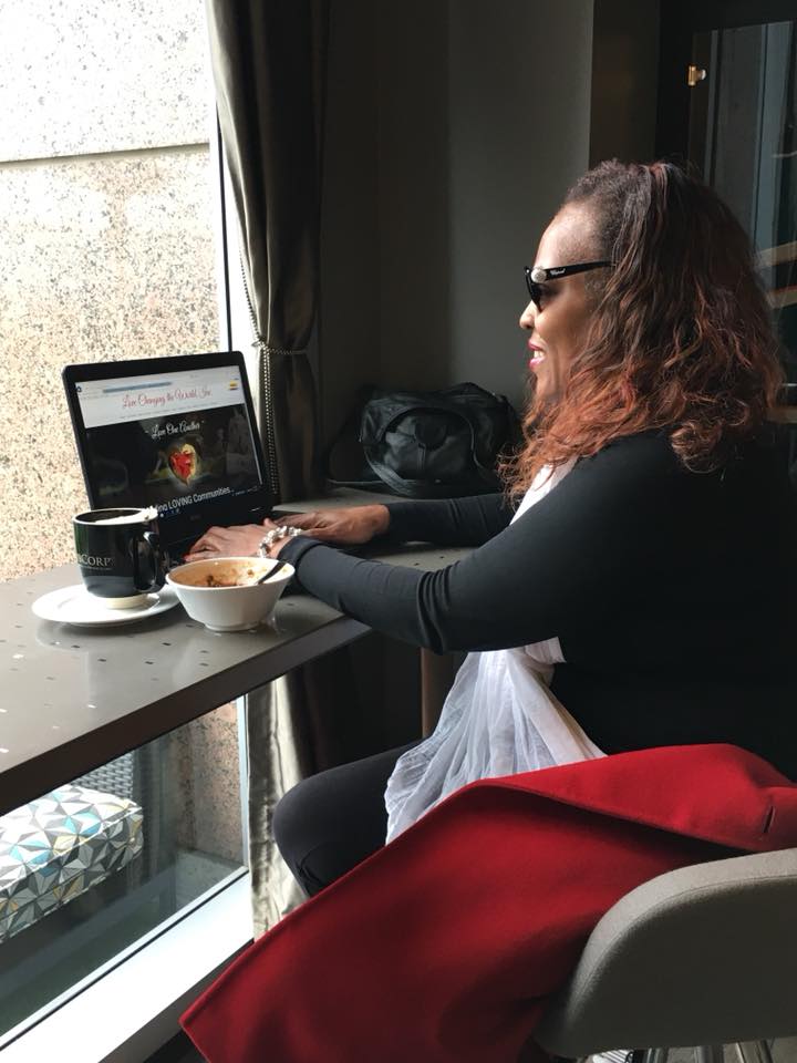 Lady sitting at a table in front of a window. She is drinking tea and working on a laptop.
