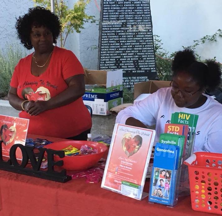 Two women representing Love Changing The World, Inc. sit at a community outreach table with educational materials about HIV and sexually transmitted infections, along with candy and prevention resources displayed for public health distribution.