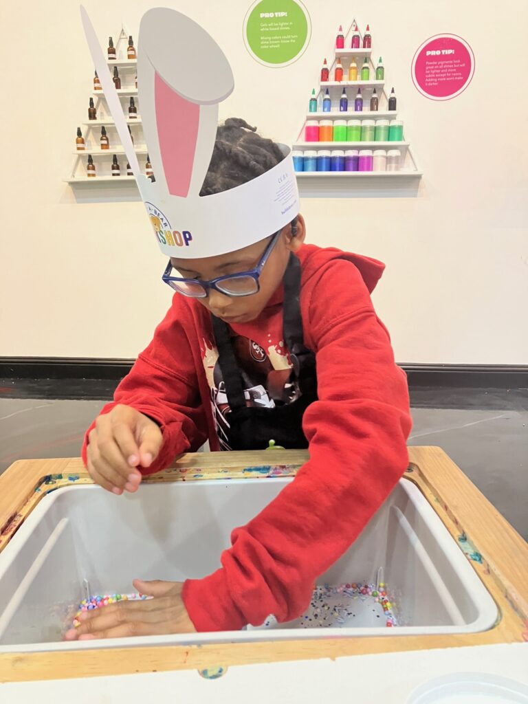 A young child wearing glasses, a red hoodie, and a paper bunny-ear hat concentrates while reaching into a craft station filled with small colorful beads. The child wears a black apron and stands at a wooden activity table, with shelves of brightly colored craft materials arranged on the wall behind them.
